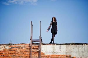 portrait fille brune aux lèvres rouges vêtue d'une robe noire posée sur le toit près de l'échelle. mannequin de rue. photo