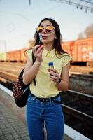 jeune adolescente debout sur le quai de la gare et soufflant des bulles de savon, portant un t-shirt jaune, un jean et des lunettes de soleil, avec sac à dos.