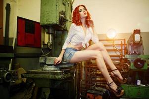 fille aux cheveux rouges portant un short en jean court et un chemisier blanc posé sur une machine industrielle à l'usine. photo