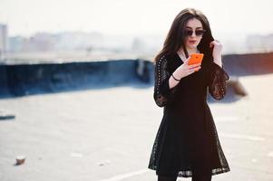 portrait fille brune aux lèvres rouges et téléphone portable orange à portée de main, vêtue d'une robe noire, lunettes de soleil posées sur le toit. mannequin de rue. photo