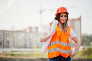 ingénieur constructeur femme en gilet uniforme et casque de protection orange tenir un papier commercial contre de nouveaux bâtiments avec grue. photo