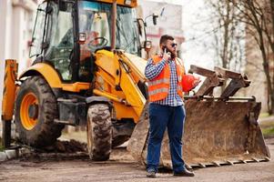 barbe travailleur homme costume travailleur de la construction en sécurité casque orange, lunettes de soleil contre tracteur avec téléphone portable à portée de main. photo