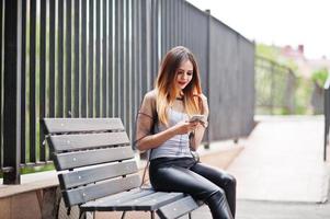 femme à la mode regarde une chemise blanche, des vêtements transparents noirs, un pantalon en cuir, posant dans la rue et assis sur un banc avec un téléphone portable. concept de fille de mode. photo