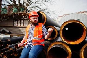 travailleur de barbe brutal homme costume travailleur de la construction dans un casque orange de sécurité près de tuyaux en acier avec un ruban à mesurer à portée de main. photo