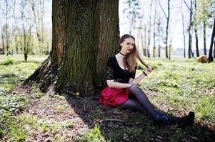portrait d'une fille avec un maquillage brillant avec des lèvres rouges, un collier tour de cou noir sur son cou et une jupe en cuir rouge assis près d'un arbre au parc. photo