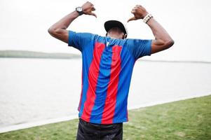 un garçon afro-américain élégant porte une casquette, un t-shirt de football montrant son dos contre le lac. portrait d'homme sportif noir. photo