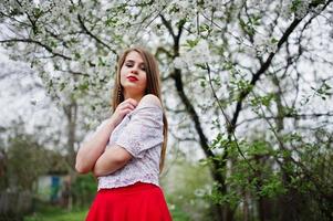 portrait de belle fille aux lèvres rouges au jardin de fleurs de printemps, porter une robe rouge et un chemisier blanc. photo