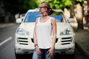femme blonde élégante portant un jean, des lunettes, un tour de cou et une chemise blanche contre une voiture de luxe. portrait de modèle urbain de mode. photo