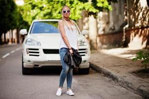 femme blonde élégante portant un jean, des lunettes de soleil, un tour de cou et une chemise blanche contre une voiture de luxe. portrait de modèle urbain de mode. photo