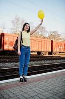 jeune adolescente debout sur le quai de la gare avec ballon à portée de main, porter sur t-shirt jaune, jeans et lunettes de soleil, avec sac à dos. photo