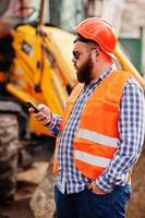 barbe travailleur homme costume travailleur de la construction en sécurité casque orange, lunettes de soleil contre tracteur avec téléphone portable à portée de main.