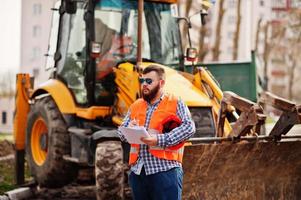 travailleur de barbe brutal homme costume travailleur de la construction dans un casque orange de sécurité, lunettes de soleil contre traktor avec plan papier à portée de main. photo