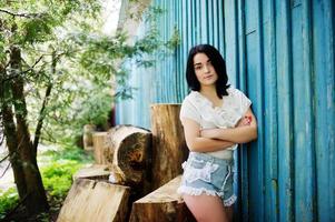 portrait d'une fille brune sexy sur un short en jean pour femme et un chemisier blanc contre une maison en bois bleue avec des souches. photo