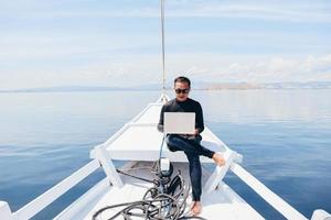 homme asiatique en combinaison et lunettes de soleil assis sur le bateau et utilisant un ordinateur portable en vacances à labuan bajo photo