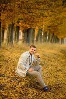 portrait d'un jeune homme en manteau sur fond d'arbres d'automne. un homme est assis par terre. photo