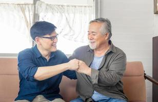 joyeux père asiatique senior et fils d'âge moyen se cognent les poings, papa d'âge mûr et homme du millénaire assis sur un canapé dans le salon à la maison, concepts de famille asiatique de bonheur photo