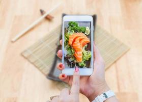 main femme avec smartphone prenant une photo de saumon sashimi avec légumes et radis en plaque noire et baguettes sur table en bois.