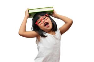 drôle et heureuse petite fille préscolaire asiatique portant des lunettes rouges tenant un livre vert sur la tête, sur fond blanc isolé. concept d'écolier et d'éducation au primaire et au préscolaire photo