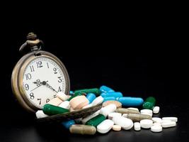 horloge antique sur multicolore de médicament et capsule est sur le fond noir. fermer. nous sommes contre les médicaments anti-médicaments, cure dans un récipient pour la santé. tas de pilules à capsules rondes blanches bleues vertes photo