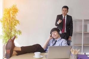 un travailleur masculin asiatique écoute de la musique avec ses pieds sur le bureau du bureau. et le superviseur se tenait avec un visage sérieux derrière lui photo