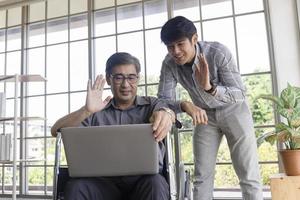 un père asiatique d'âge moyen est assis dans un fauteuil roulant et a un fils à côté de lui qui discute sur des appels vidéo sur un ordinateur portable. photo