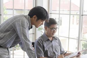 un homme asiatique d'âge moyen enseignant à son fils un travail debout à côté d'un bureau dans un bureau. photo