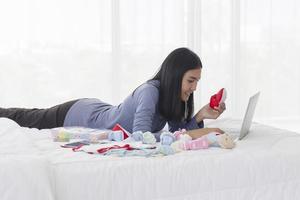 une mère asiatique achète des vêtements de bébé pour son enfant sur un ordinateur portable, fait des achats en ligne au lit avec un sourire radieux. photo