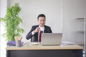 un gestionnaire asiatique travaillant dans un bureau tenant une tasse de café blanche avec un sourire rayonnant. photo