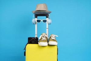 valise jaune avec lunettes de soleil, chapeau et appareil photo sur fond bleu pastel. notion de voyage. style minimaliste