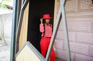 jolies tresses affaires femme afro-américaine brillante personne autoritaire amicale porter bureau chemise rouge, chapeau et pantalon. photo