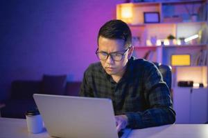 homme indépendant travaillant sur ordinateur portable à la maison la nuit. programmeur développeur homme, ingénieur de données, pensant, sérieux tout en travaillant à la maison la nuit. travail indépendant et concept de travail à domicile. photo