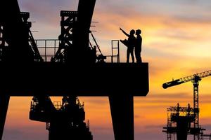 silhouette d'ingénieur et de travailleur vérifiant le projet sur fond de chantier d'infrastructure lourde, chantier de construction au coucher du soleil en soirée. photo