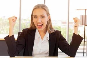 une belle femme d'affaires en costume, bien habillée, assise au bureau, heureuse avec enthousiasme, leva la main avec un sourire éclatant sur la table photo