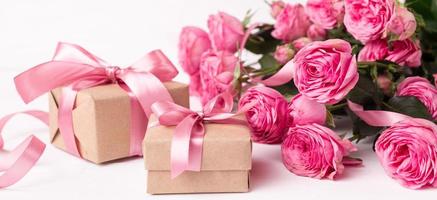 des roses rose pastel fraîches et des coffrets cadeaux enveloppés dans du papier kraft avec des rubans sur une table en bois blanche.