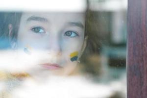 un enfant triste regarde la fenêtre avec le drapeau de l'ukraine peint sur la joue, les soucis et la peur. aide humanitaire aux enfants, paix mondiale, sécurité. photo