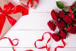 ruban rouge en forme de coeur, bouquet de roses et coffrets cadeaux enveloppés dans du papier kraft sur une table en bois blanche.