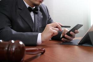 contexte de la justice et du droit. main d'avocat masculin travaillant avec un téléphone intelligent, un clavier d'accueil pour ordinateur tablette numérique avec un marteau et un document sur une table en bois photo