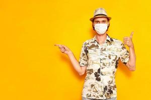 un touriste en vacances masculin en masque médical pointe son doigt sur une zone vide et se tient avec son pouce levé isolé sur fond jaune vêtu d'une chemise, d'un chapeau. concept repos, vacances, voyage photo