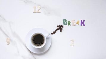 vue de dessus du café noir dans une tasse en céramique blanche et des grains de café dans une horloge en forme de main avec des chiffres en bois et un mot de rupture dans le cadran de l'horloge se formant sur une table en marbre blanc photo