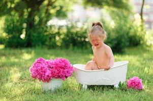 une petite fille heureuse prend un bain de lait avec des pétales. petite fille dans un bain de lait sur fond vert. bouquets de pivoines roses. bain de bébé. hygiène et soins aux jeunes enfants. photo