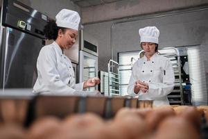 deux femmes chefs professionnelles en uniformes et tabliers de cuisinier blancs pétrissent la pâte à pâtisserie et les œufs, préparent du pain, des biscuits et des aliments de boulangerie frais, cuisant au four dans une cuisine en acier inoxydable d'un restaurant. photo