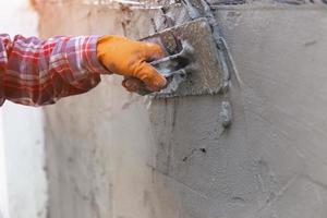 les mains d'un ouvrier qui plâtre de près portent des gants en caoutchouc orange pour empêcher le ciment de se mordre les mains, construisant les murs de la maison et ayant de belles lumières orange. photo