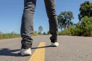 pieds d'homme debout sur une route goudronnée avec des lignes de marquage jaunes. homme portant des baskets et des jeans. photo