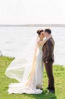 la mariée et le marié s'embrassent près de l'étang dans la nature. le vent soulève un léger voile.