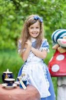 une petite fille mignonne dans le costume d'alice du pays des merveilles organise un goûter à sa table magique. photographié dans la nature. photo