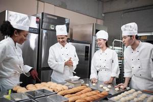 équipe gastronomique professionnelle multiraciale, quatre chefs en uniformes et tabliers de cuisinier blancs pétrissent la pâte à pâtisserie et la farine, préparent le pain et les aliments de boulangerie, cuisent au four dans la cuisine du restaurant en acier inoxydable. photo