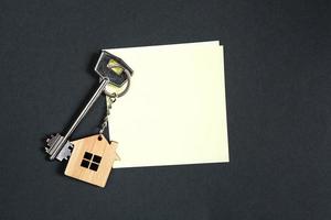 porte-clés en forme de maison en bois avec clé sur fond noir avec une feuille carrée pour les notes. construction, conception, projet, déménagement dans une nouvelle maison, hypothèque, location et achat immobilier. copie espace photo