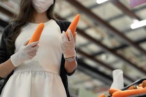 femme portant un masque protecteur et un gant en caoutchouc tenant une carotte dans un grand magasin d'épicerie de légumes. achats au supermarché dans un nouveau concept de mode de vie normal pendant la pandémie de coronavirus ou de covid. photo