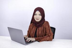 jeune femme islamique asiatique est assise et sourit en travaillant sur un ordinateur portable sur fond blanc. photo