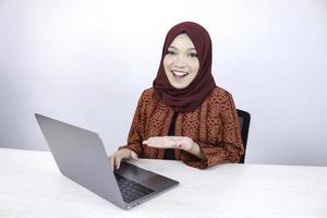 jeune femme islamique asiatique sourit en pointant la main lorsqu'elle travaille sur un ordinateur portable sur fond blanc. photo